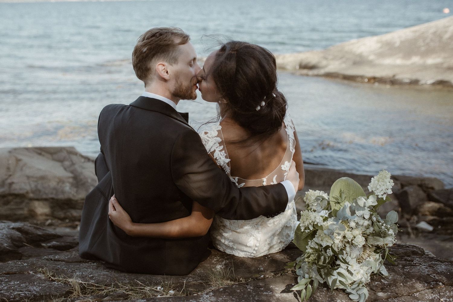 Intimate moment between newlyweds sitting on rocky coastline with bouquet.