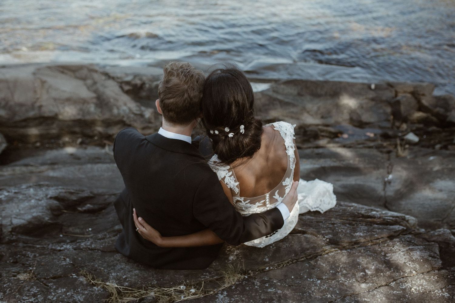Couple sits together on rocky shoreline overlooking water during sunset.