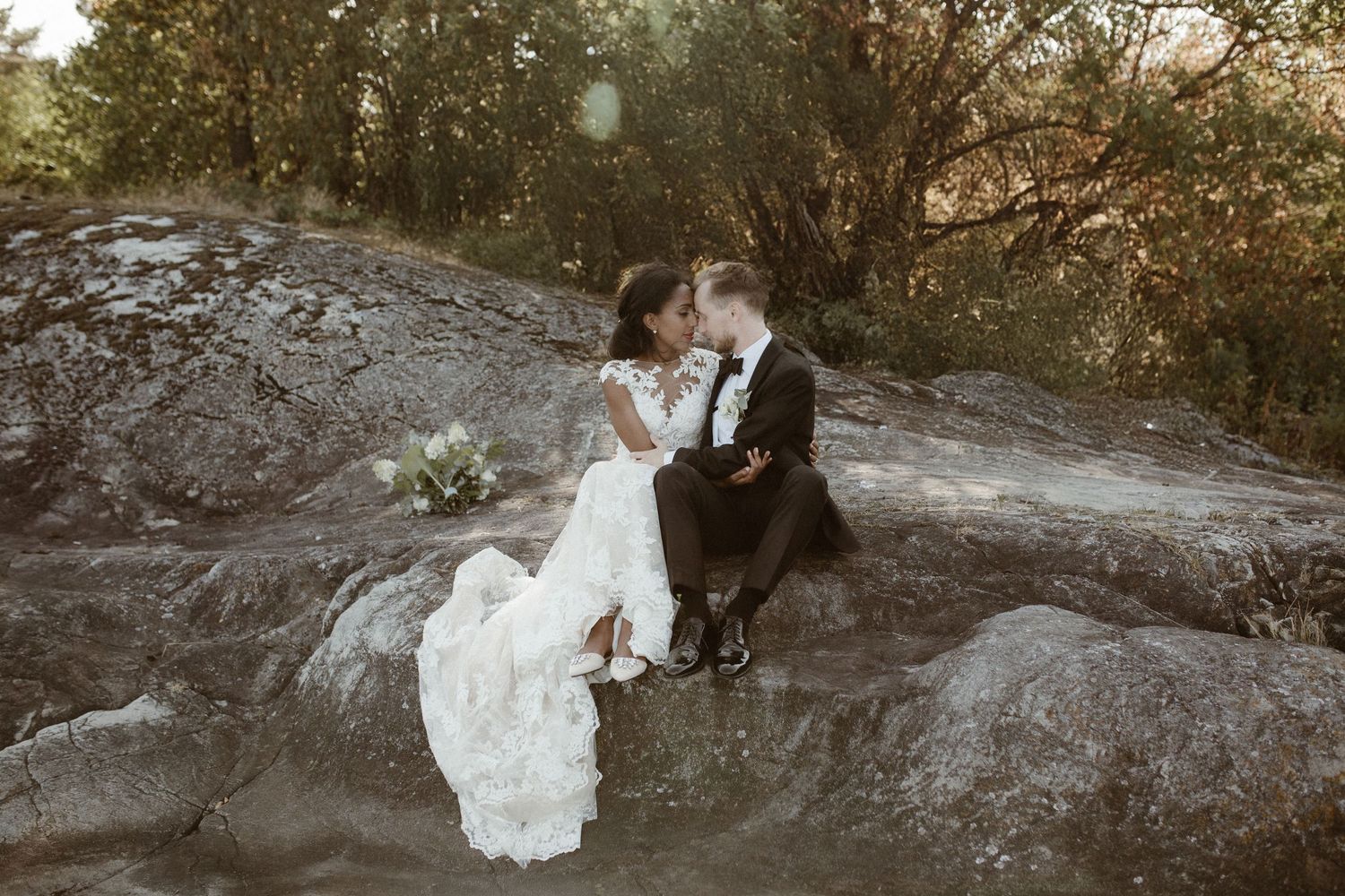 Newlyweds share intimate moment while seated on dramatic rocky outcrop at sunset.