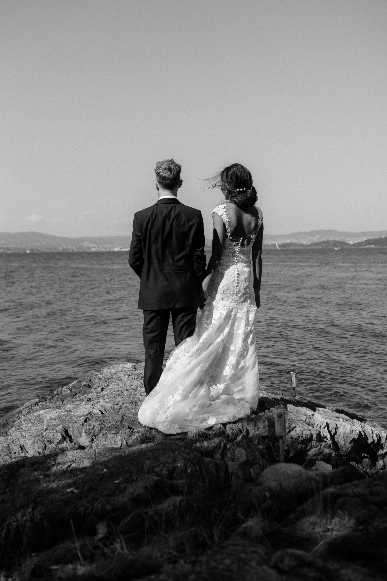 A black and white photo of a couple in wedding attire overlooking the water from rocky cliffs.