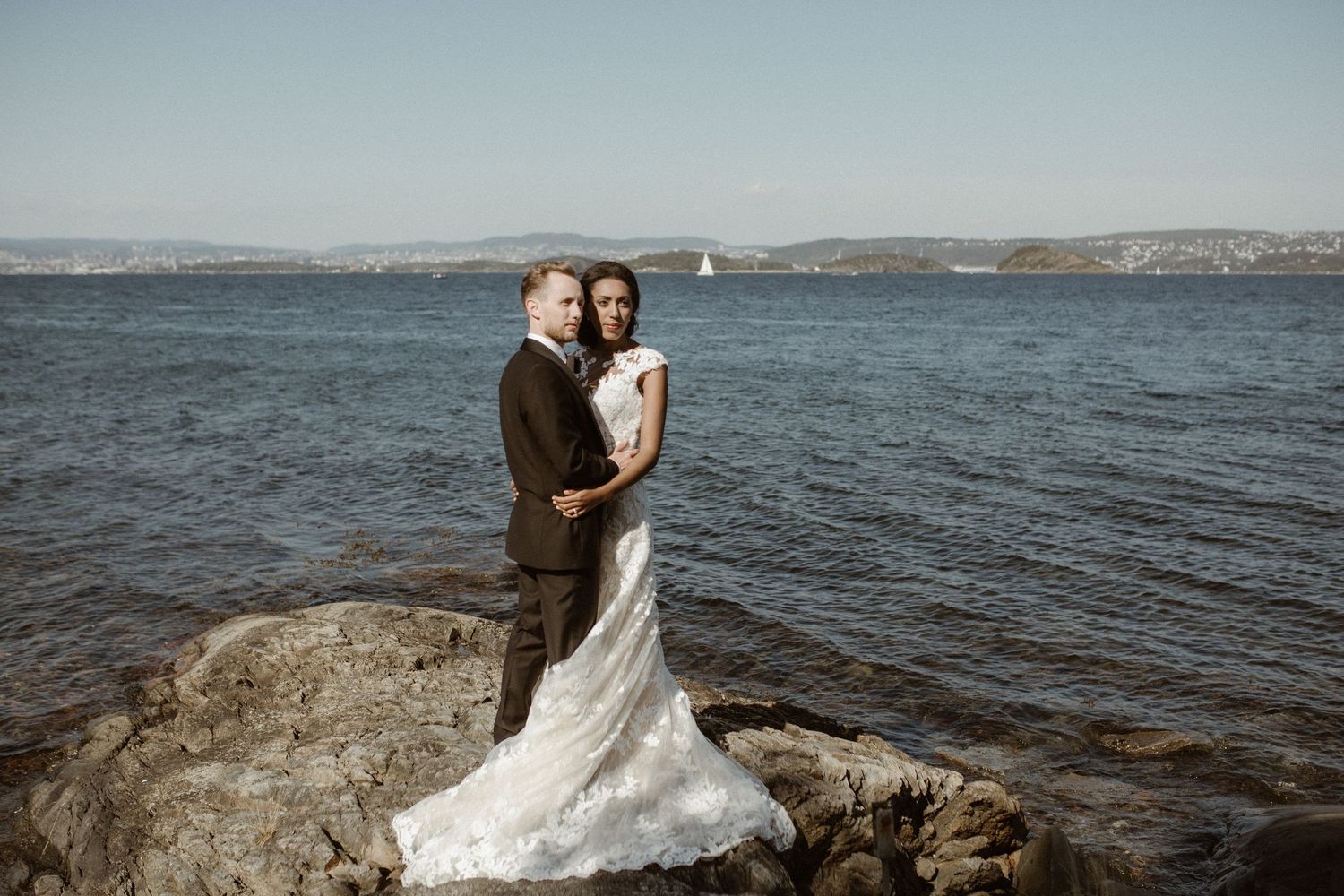 A couple in wedding attire embraces on coastal rocks with ocean and mountains in the distance.