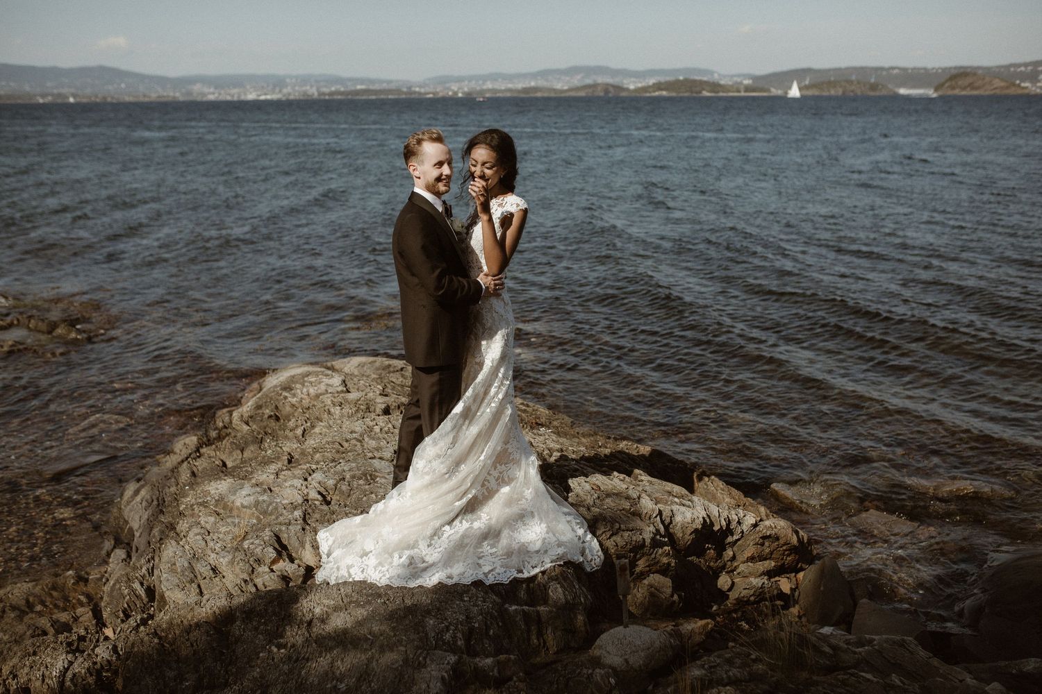 A couple stands together on a rocky outcrop overlooking a vast lake during their wedding day.