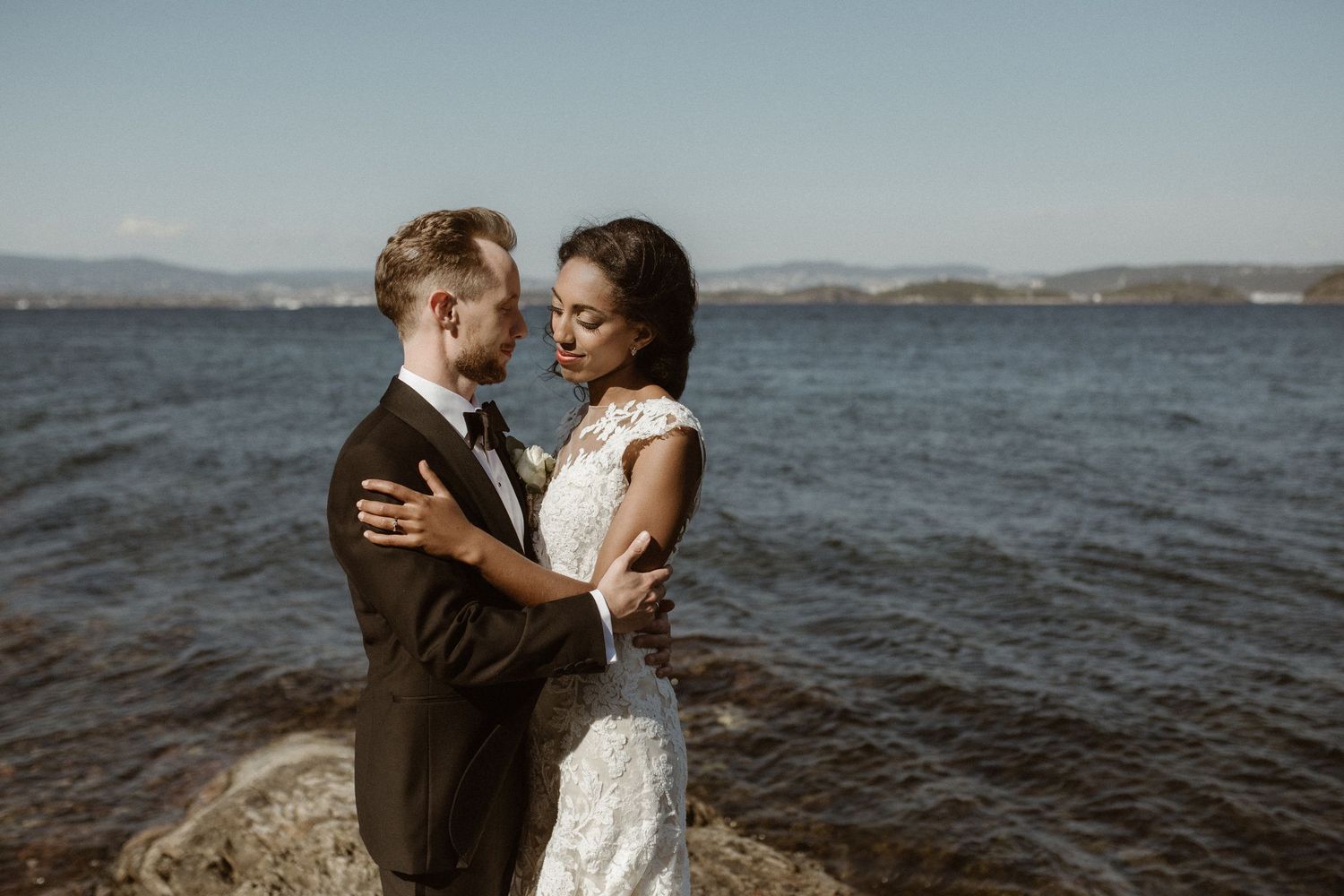 Elegant bride and groom embrace on coastal rocks with ocean and mountains in background.