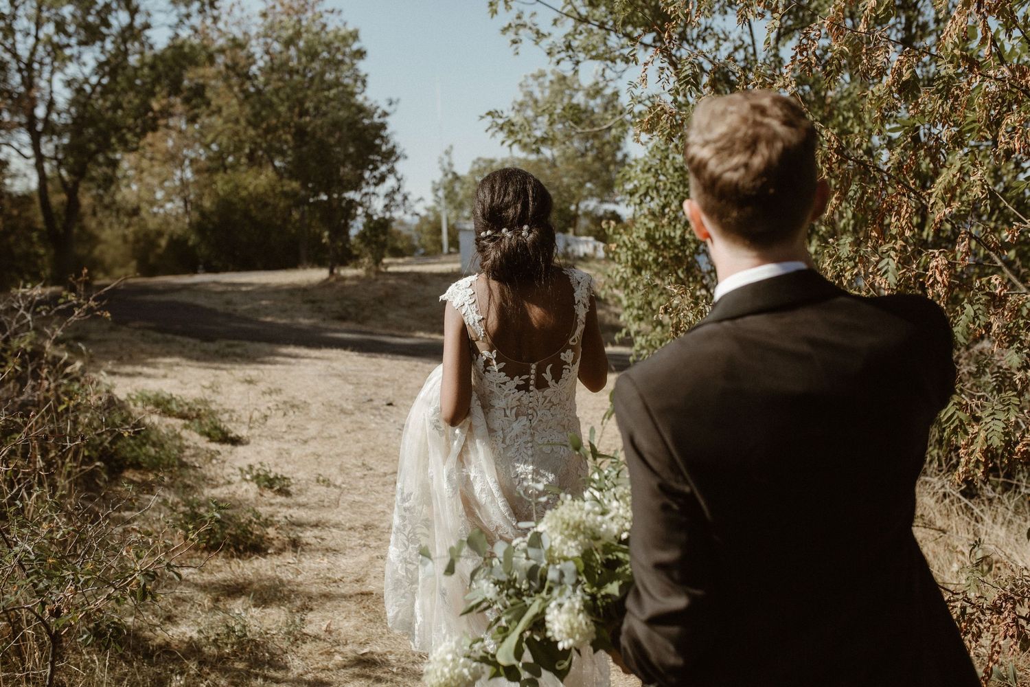 A couple walks down a sunlit trail on their wedding day, surrounded by lush foliage.
