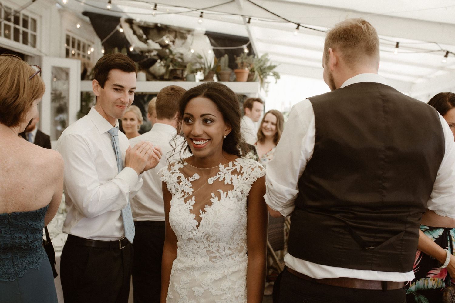 A joyful wedding reception scene shows guests dancing and celebrating under string lights in a white tent venue.