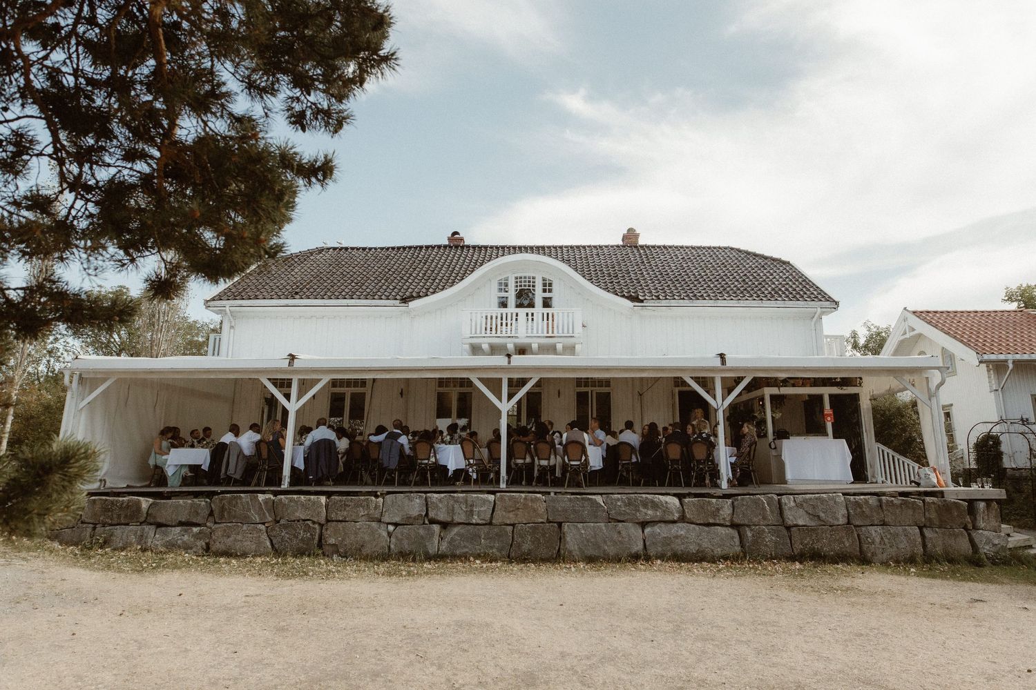 Elegant white wedding venue with wraparound porch and stone foundation hosts guests during outdoor reception.