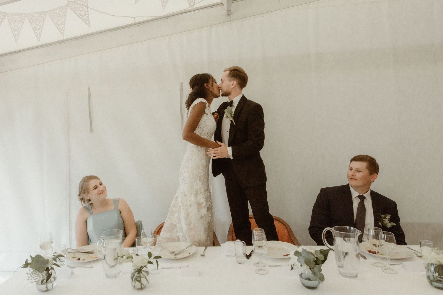 Couple sharing an intimate moment at their wedding reception head table while guests look on at decorated venue.