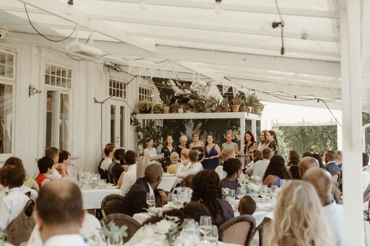 Wedding guests enjoy dinner at long tables under white-beamed ceiling during indoor reception.
