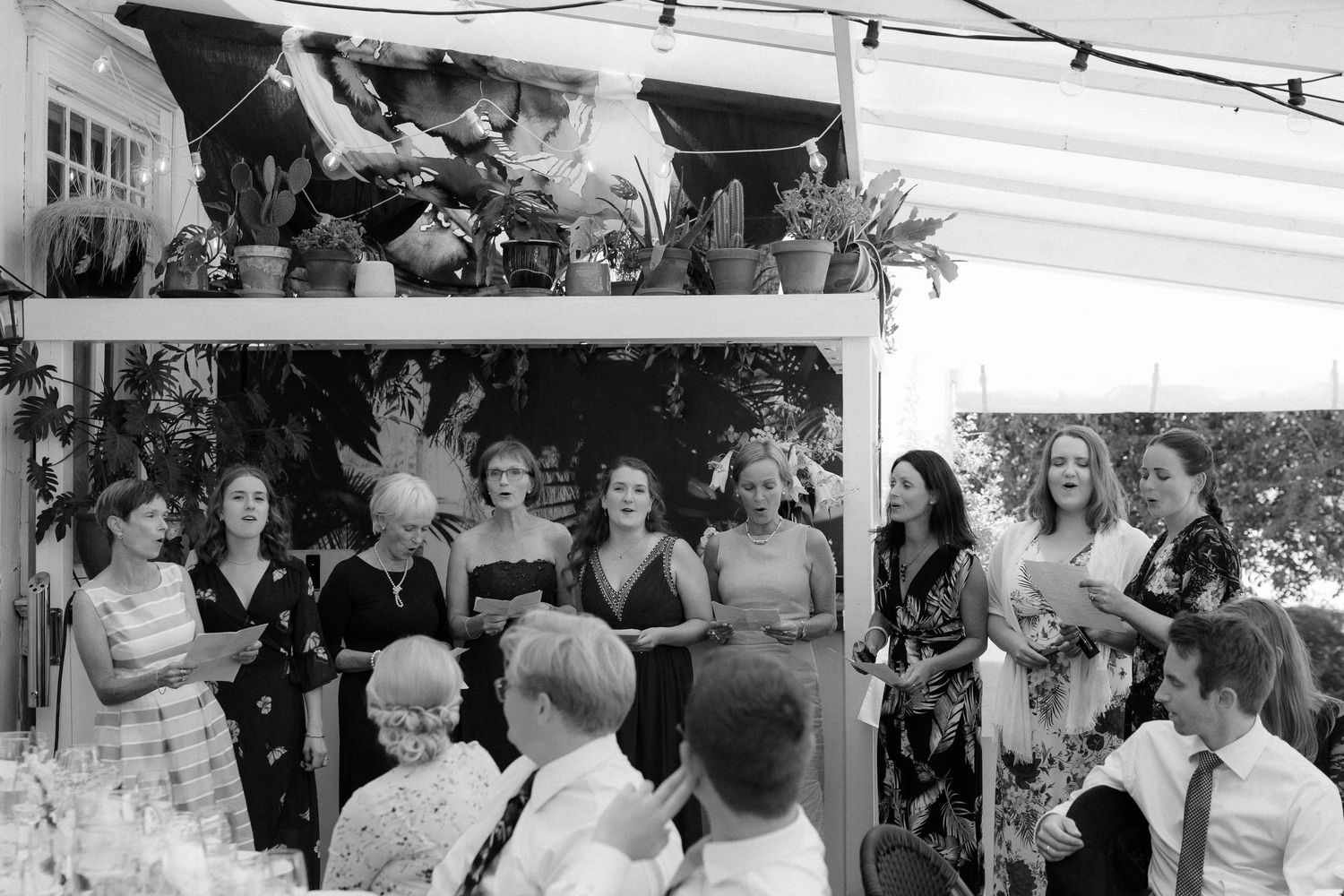 Wedding guests gather around outdoor tables during a celebratory reception with string lights overhead.