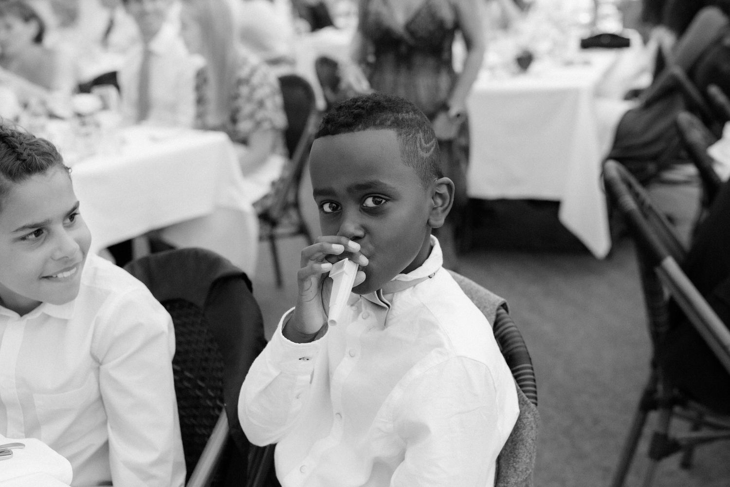 A young child in white attire at a formal event looks thoughtfully while eating.