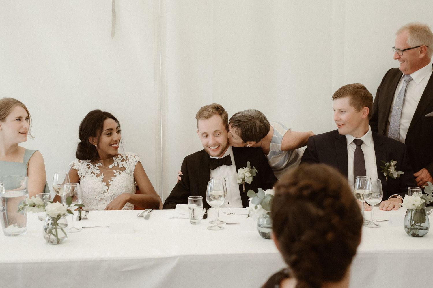 Wedding party seated at long head table during indoor reception celebration.