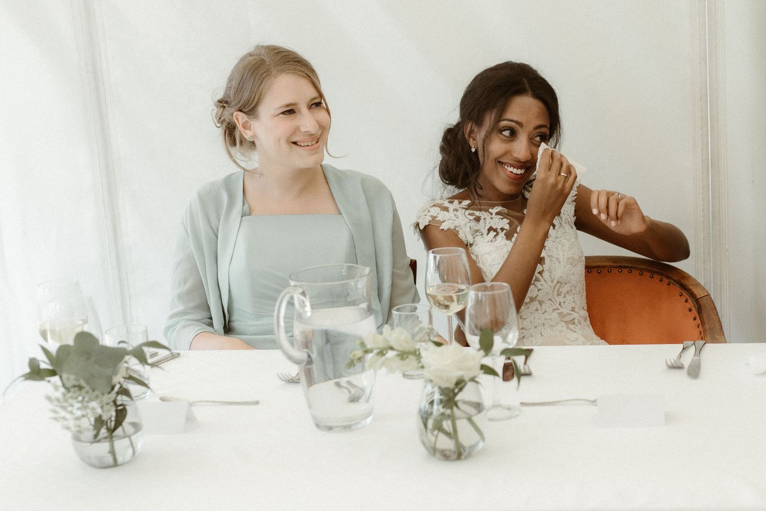 Two wedding attendees share a joyful moment while seated at an elegant table decorated with small floral arrangements.