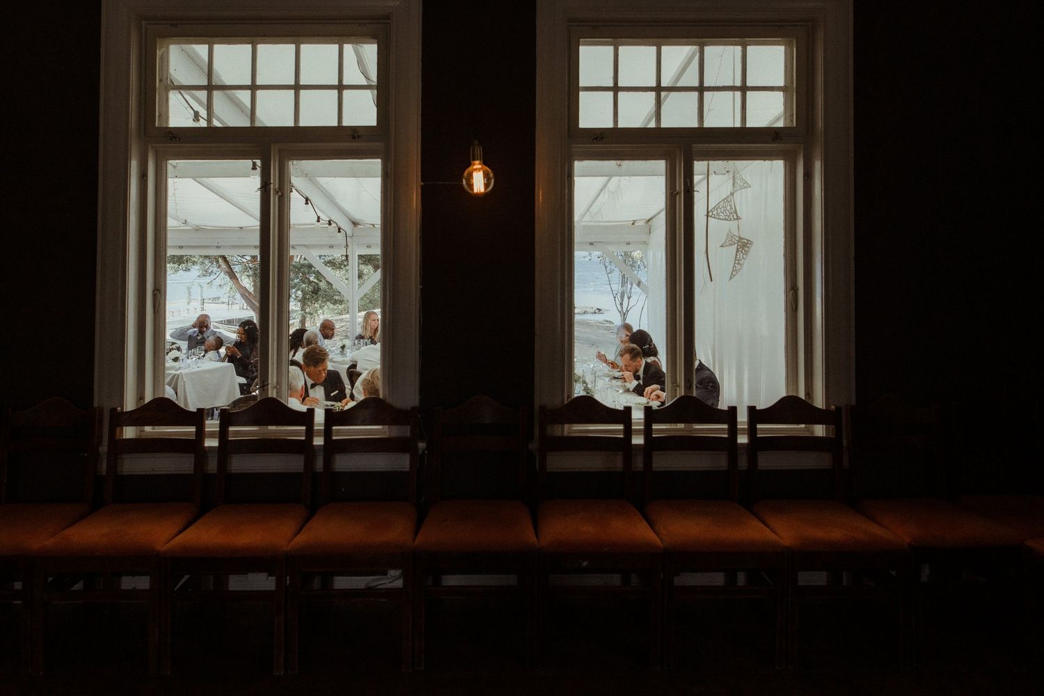 Dark window frames illuminated with warm light showing silhouettes of wedding reception guests inside.