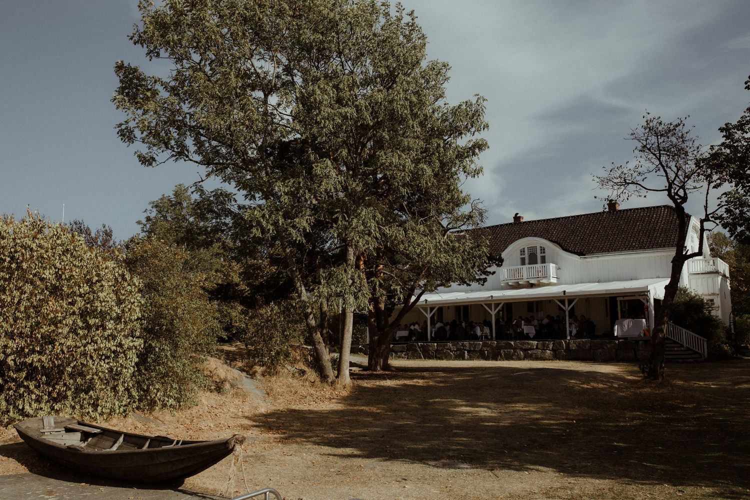 A rustic white house with wraparound porch sits among trees near a beached wooden boat.