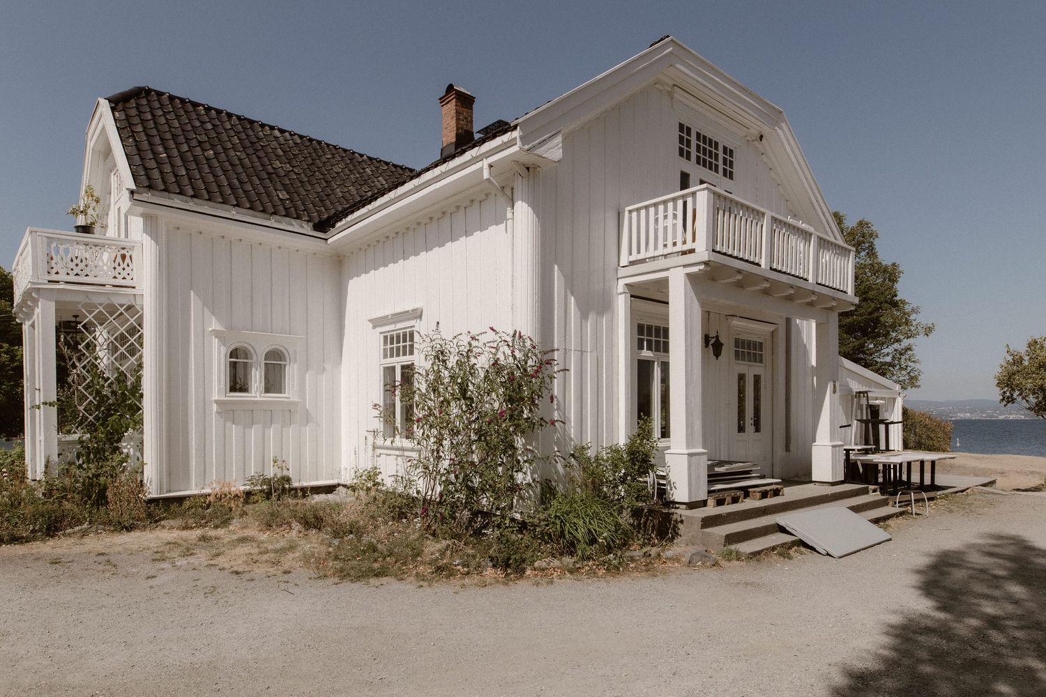 A vintage white summer house with a wrap-around porch and balcony sits along a sunny coastal shoreline.