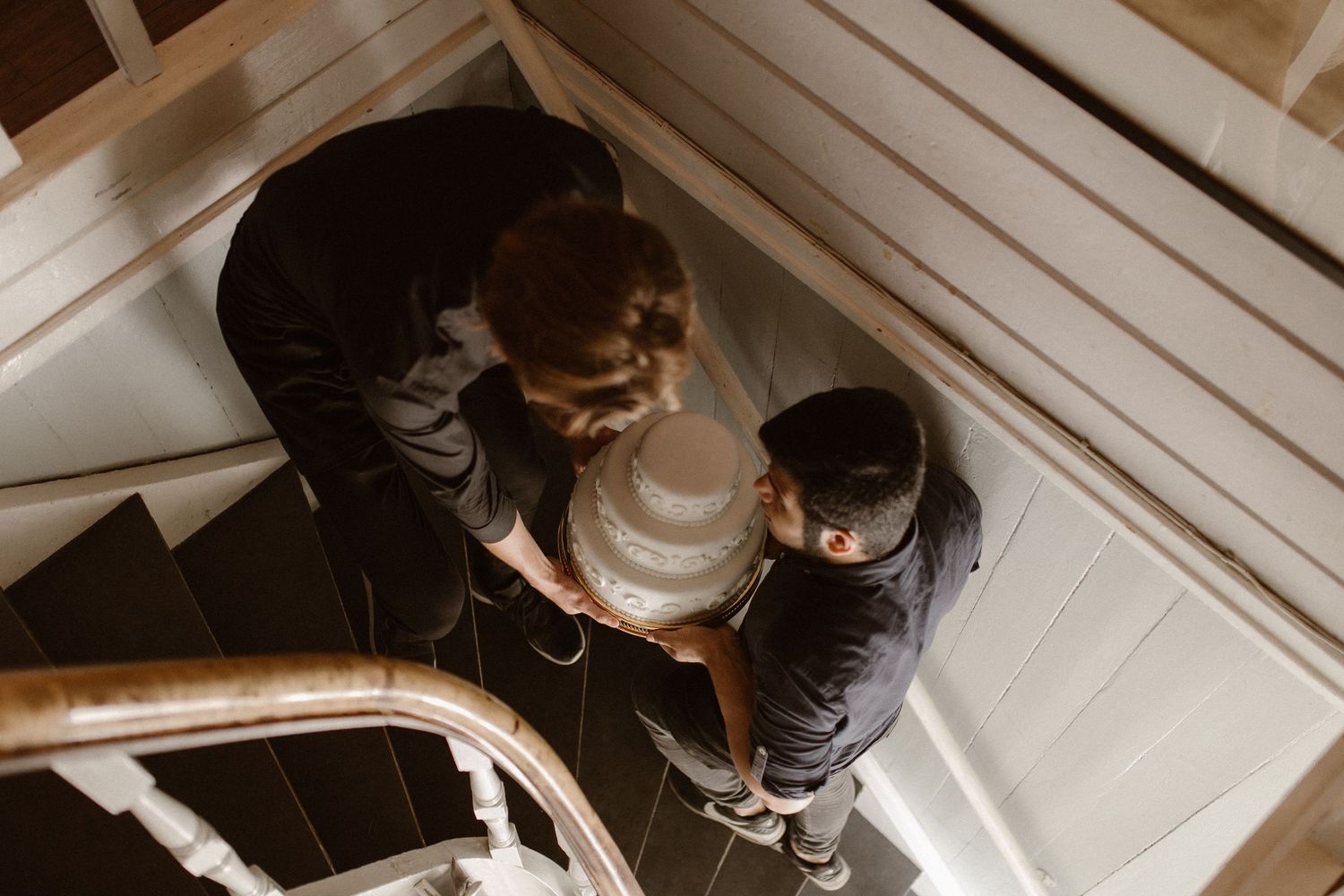 Two people stand intimately close together on white wooden stairs viewed from above in warm lighting.