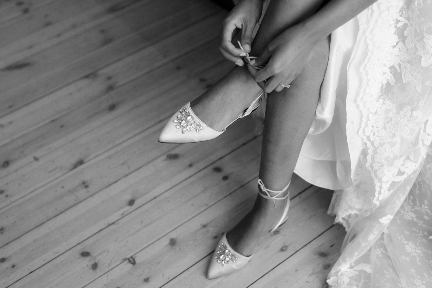 Black and white photo of someone fastening delicate lace wedding shoes on a wooden floor.