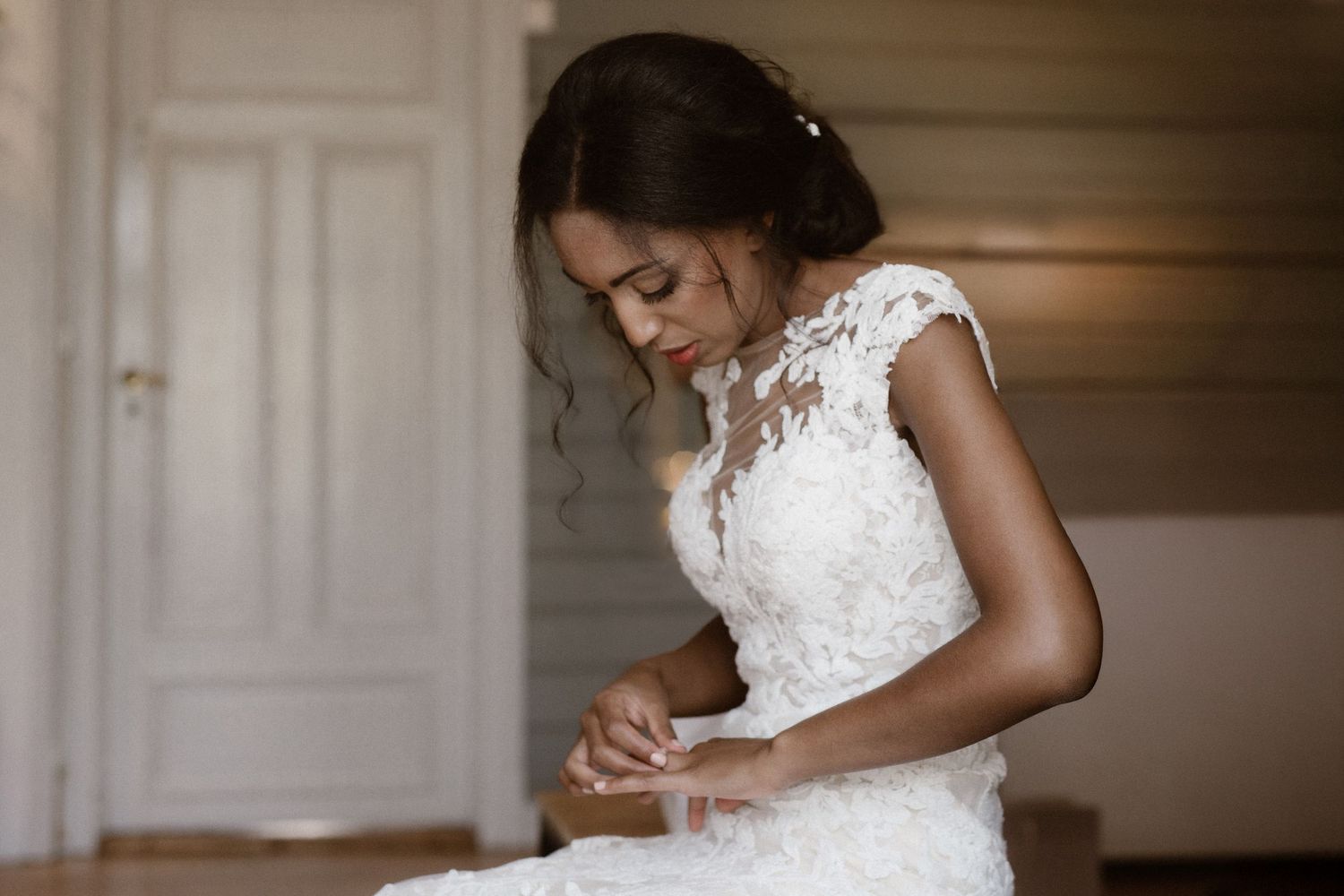 A bride in an elegant white lace wedding dress adjusts her gown while getting ready in a sunlit room.