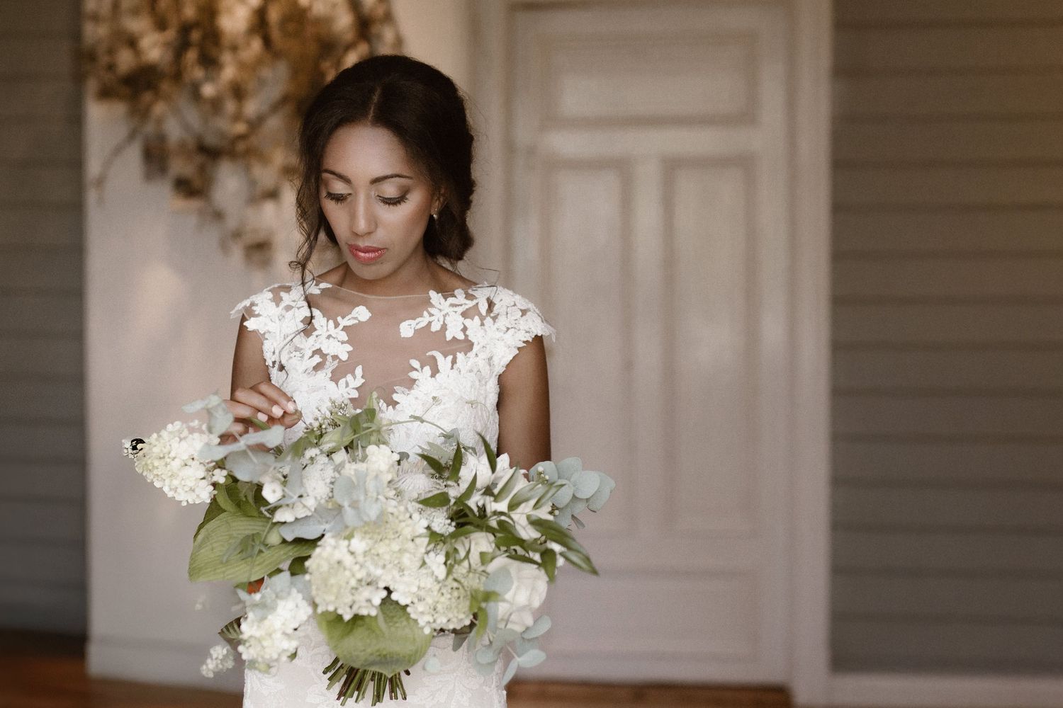 A bride in a white lace dress holds an elegant white and green floral bouquet in an elegant room with a chandelier.