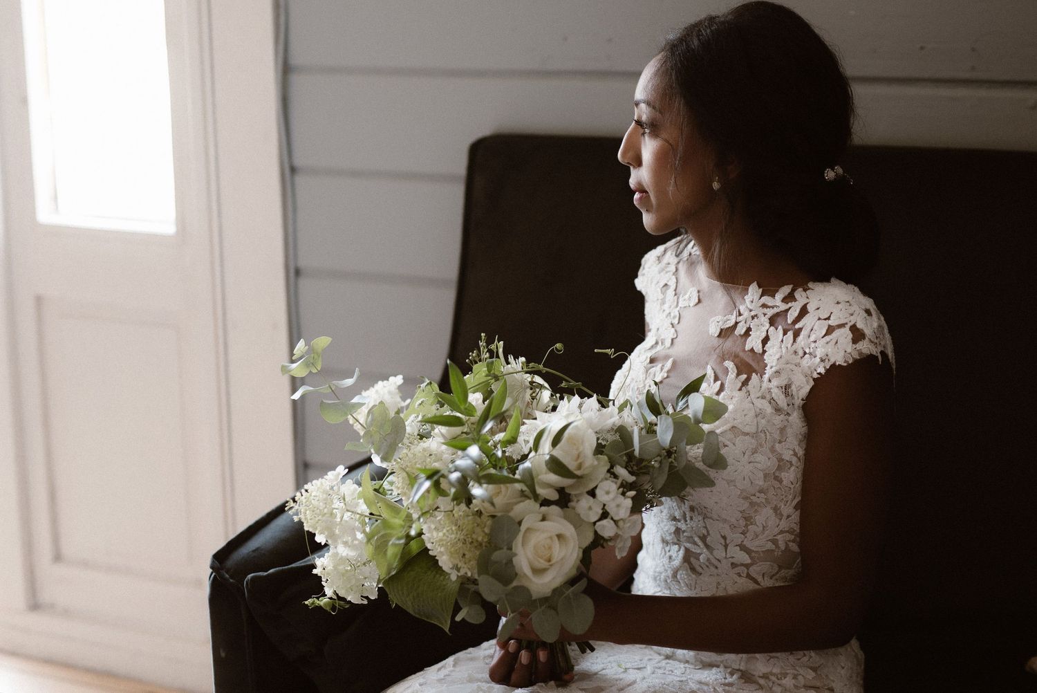 A bride wearing a delicate lace gown sits by a window holding a beautiful white and green bouquet.