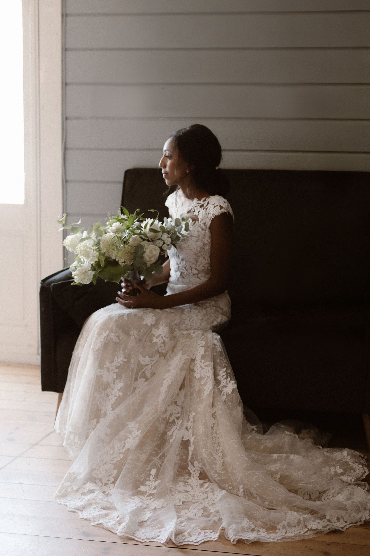 Elegant bride in lace wedding gown sits on dark couch holding white floral bouquet near window.