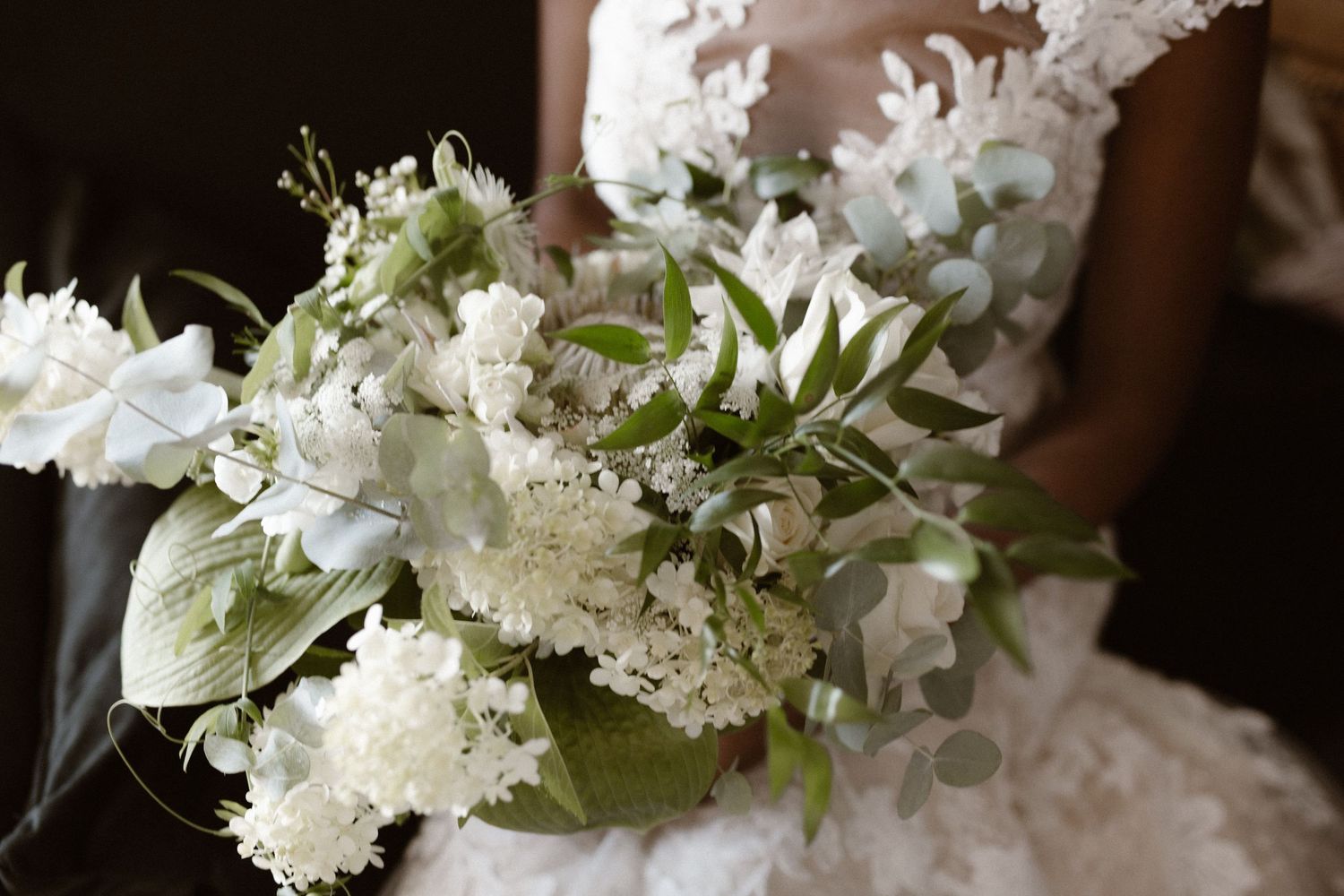 A romantic white wedding bouquet with hydrangeas and mixed greenery resting on a lace dress.