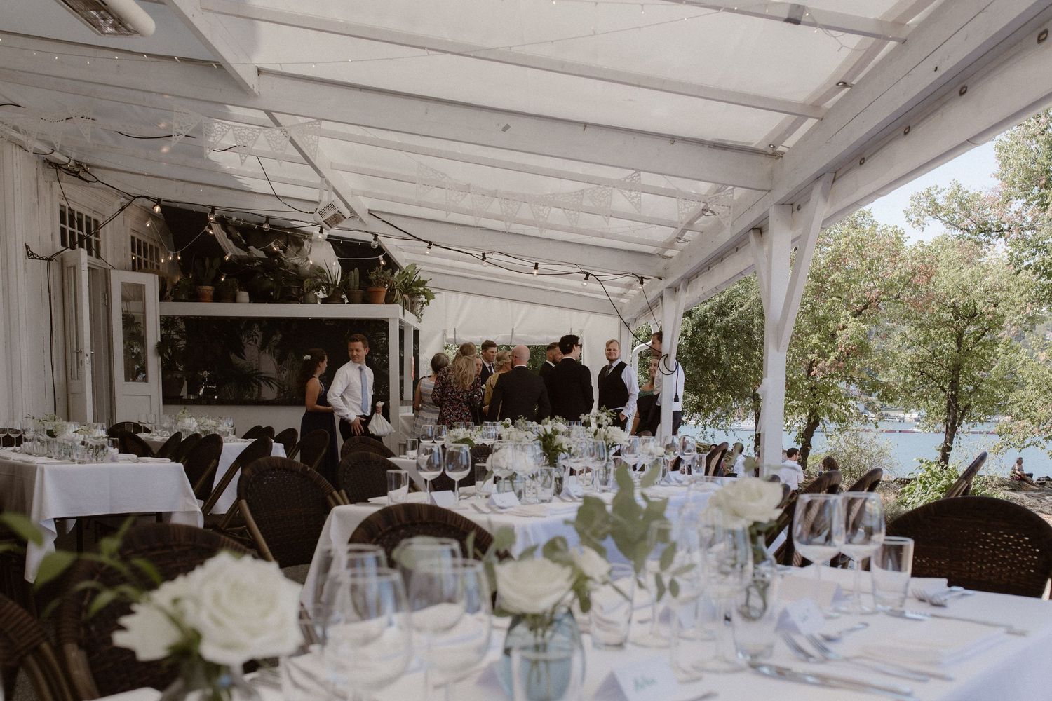 Elegant outdoor wedding reception setup with white table settings and floral centerpieces under a covered patio by the water.