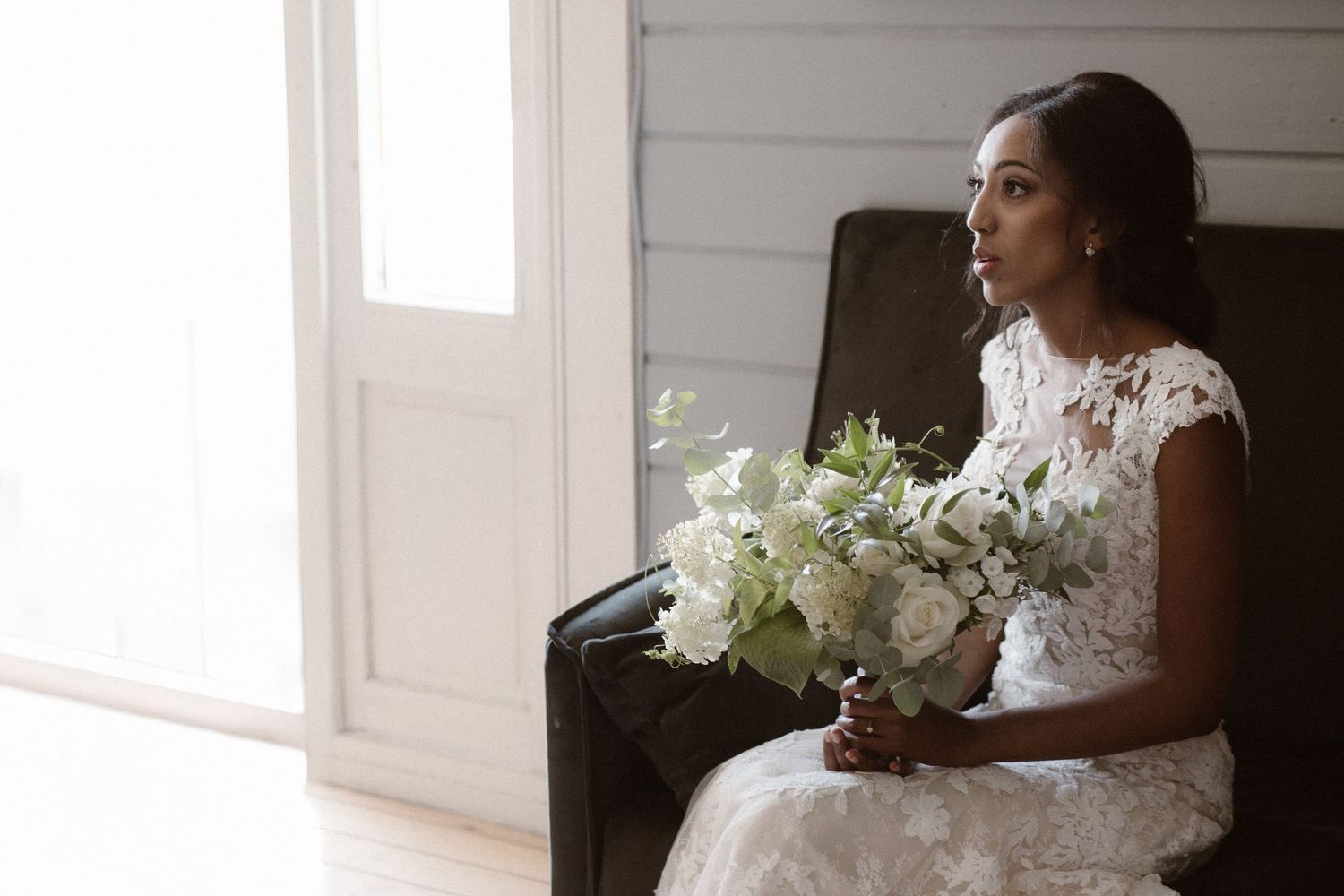 An elegant bride in a lace wedding dress sits in a chair holding a white bouquet while gazing out a nearby window.