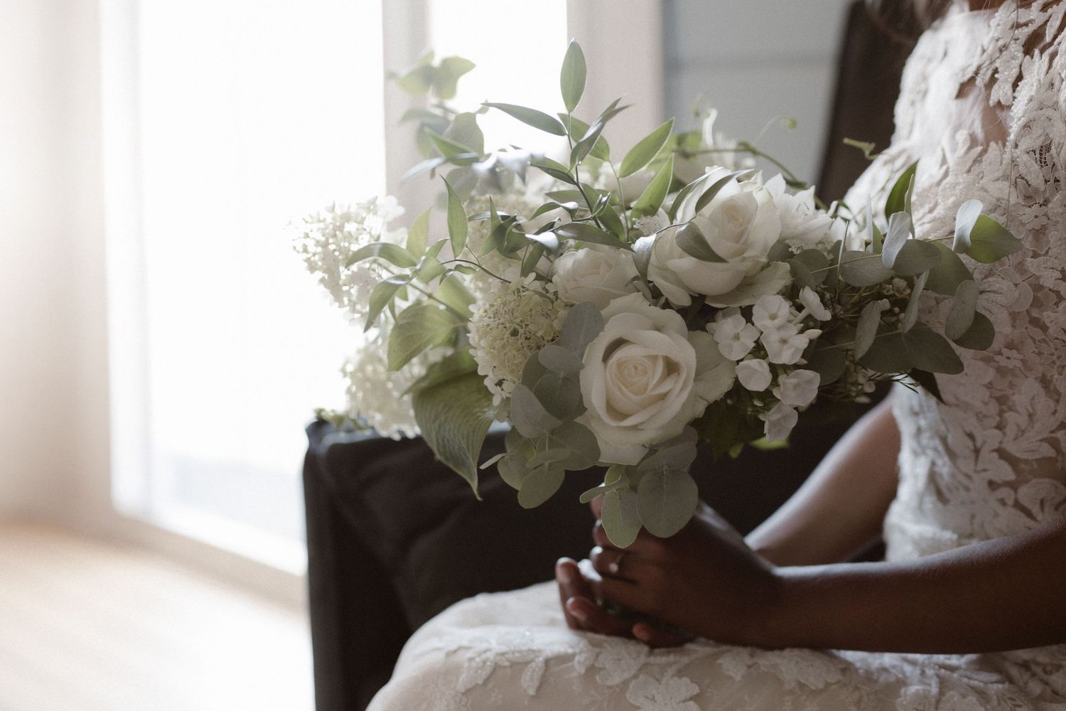 An elegant white rose bouquet with eucalyptus leaves rests on white lace fabric in natural light.