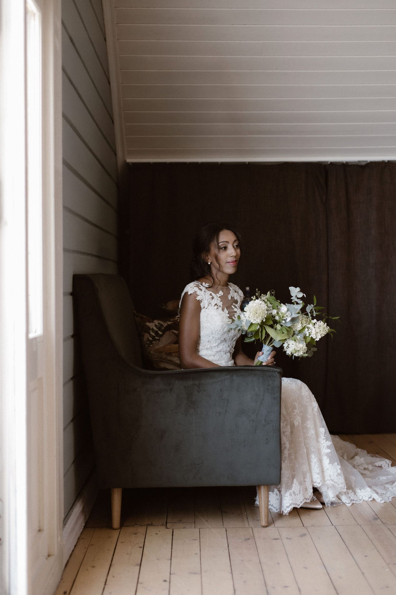 Bride in detailed lace dress poses with white bouquet against rustic wall in window light.
