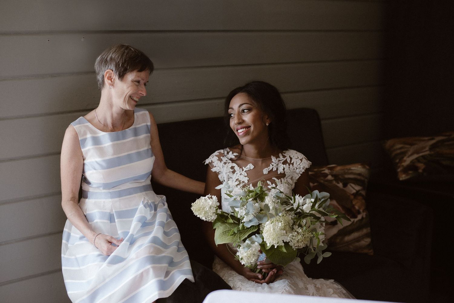 Two wedding attendees share a candid moment while sitting together against wooden wall.