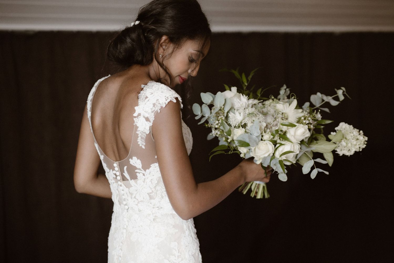 A person wearing a lace wedding dress with open back design holds a bouquet of white flowers against a dark background.