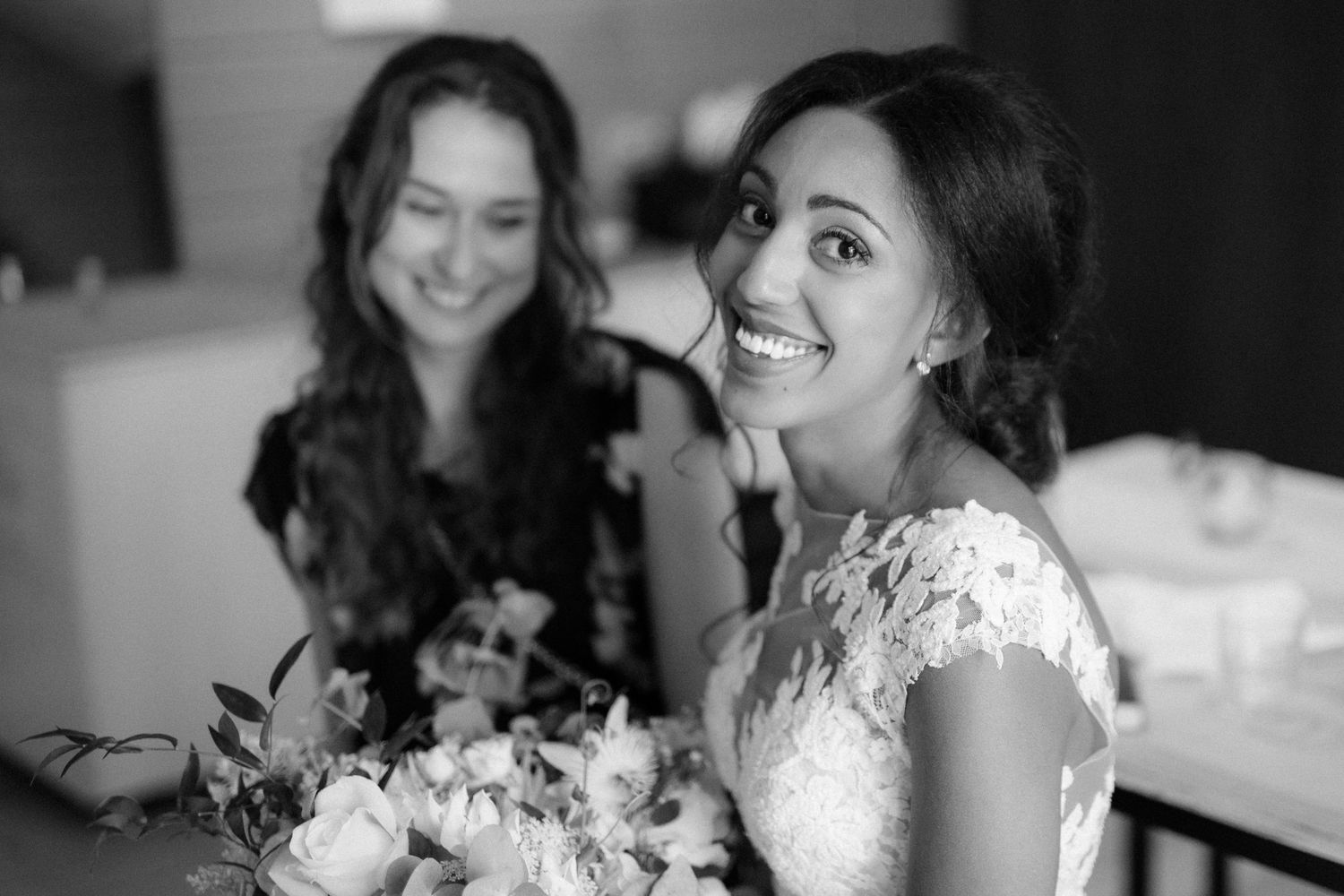 Black and white candid photograph showing joyful moments during wedding preparation with lace dress and flowers.