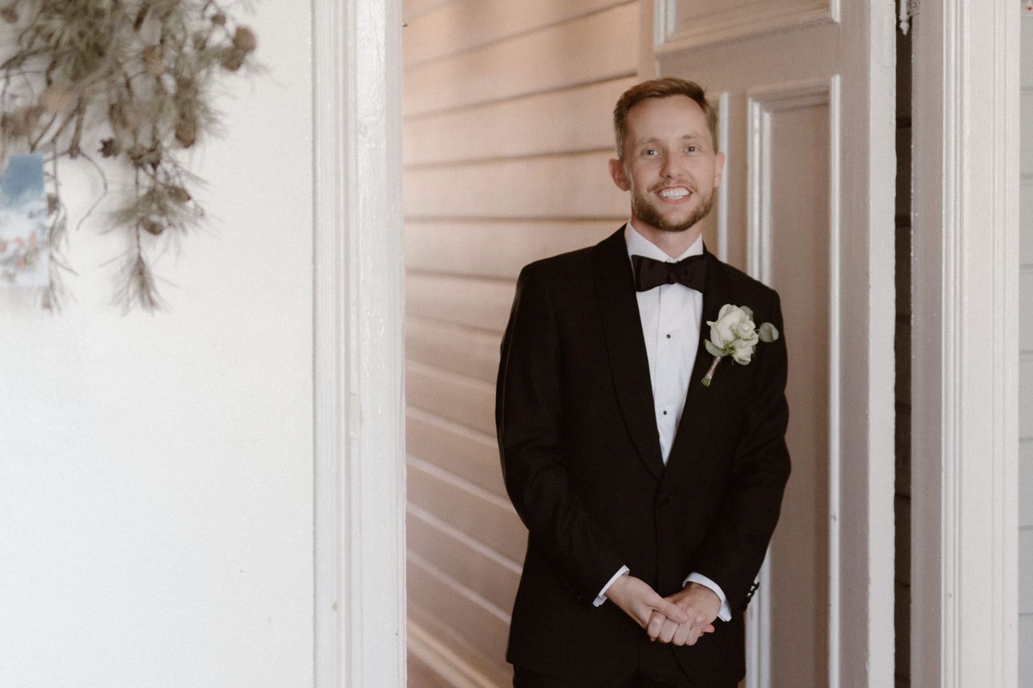 A person wearing a classic black tuxedo with bow tie and boutonniere poses against a light colored wall.