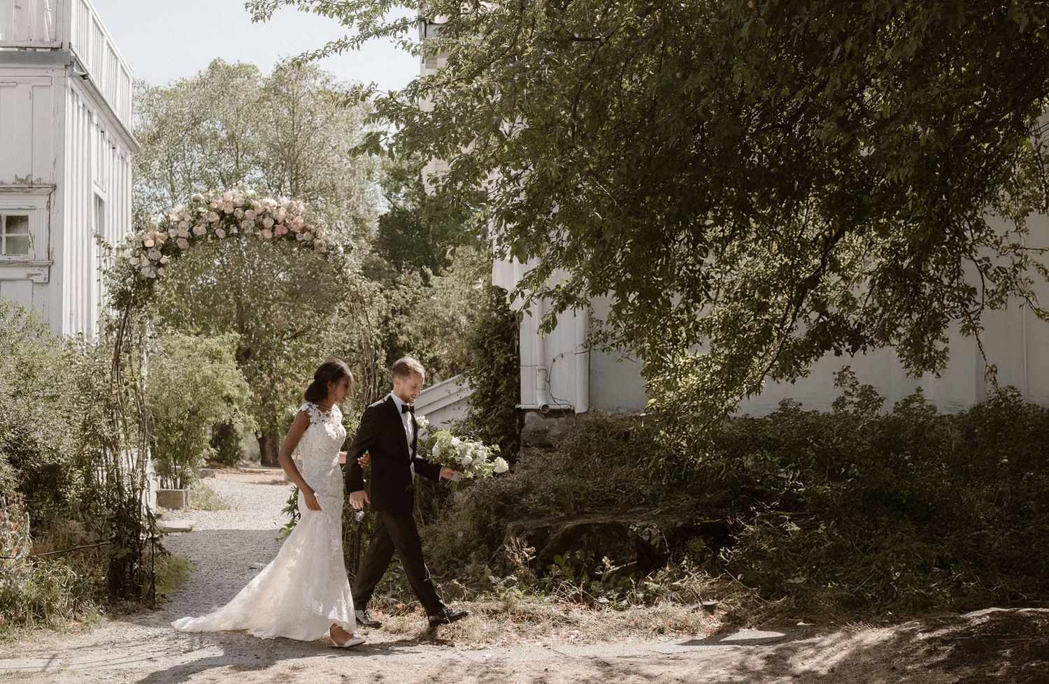 A couple in wedding attire walk together down a sunlit garden path surrounded by lush greenery and white buildings.
