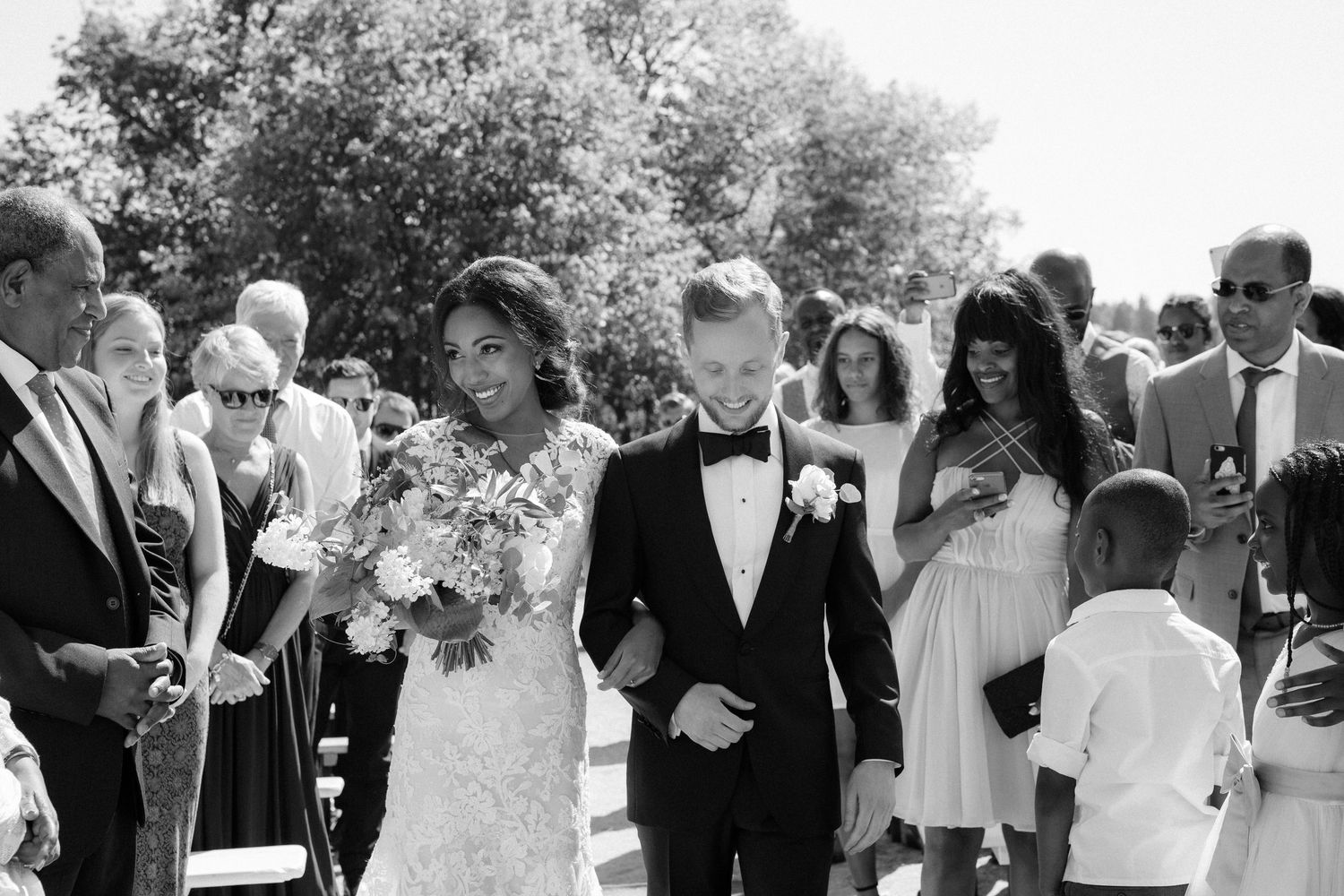 A joyful wedding party walks together outdoors on a sunny summer day in black and white photography.