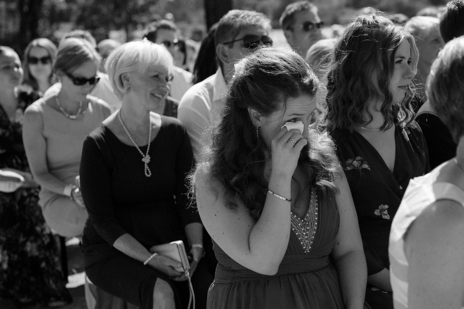 A crowd of emotional wedding guests gather outdoors during what appears to be a touching ceremony moment.