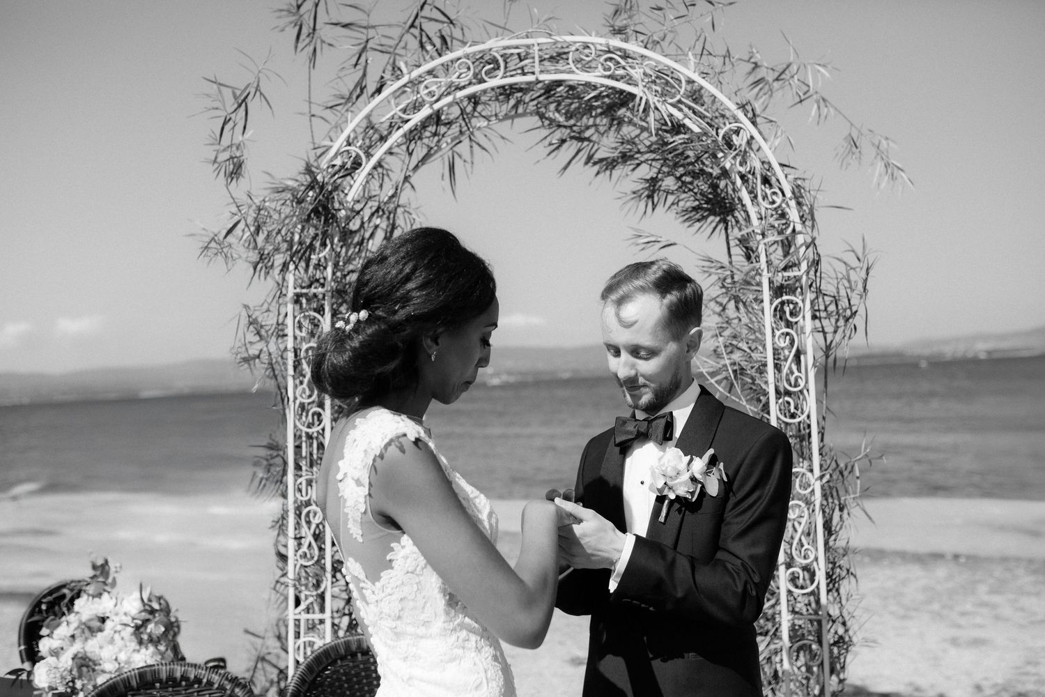 A romantic wedding ceremony takes place beneath an ornate floral arch beside the ocean in black and white.