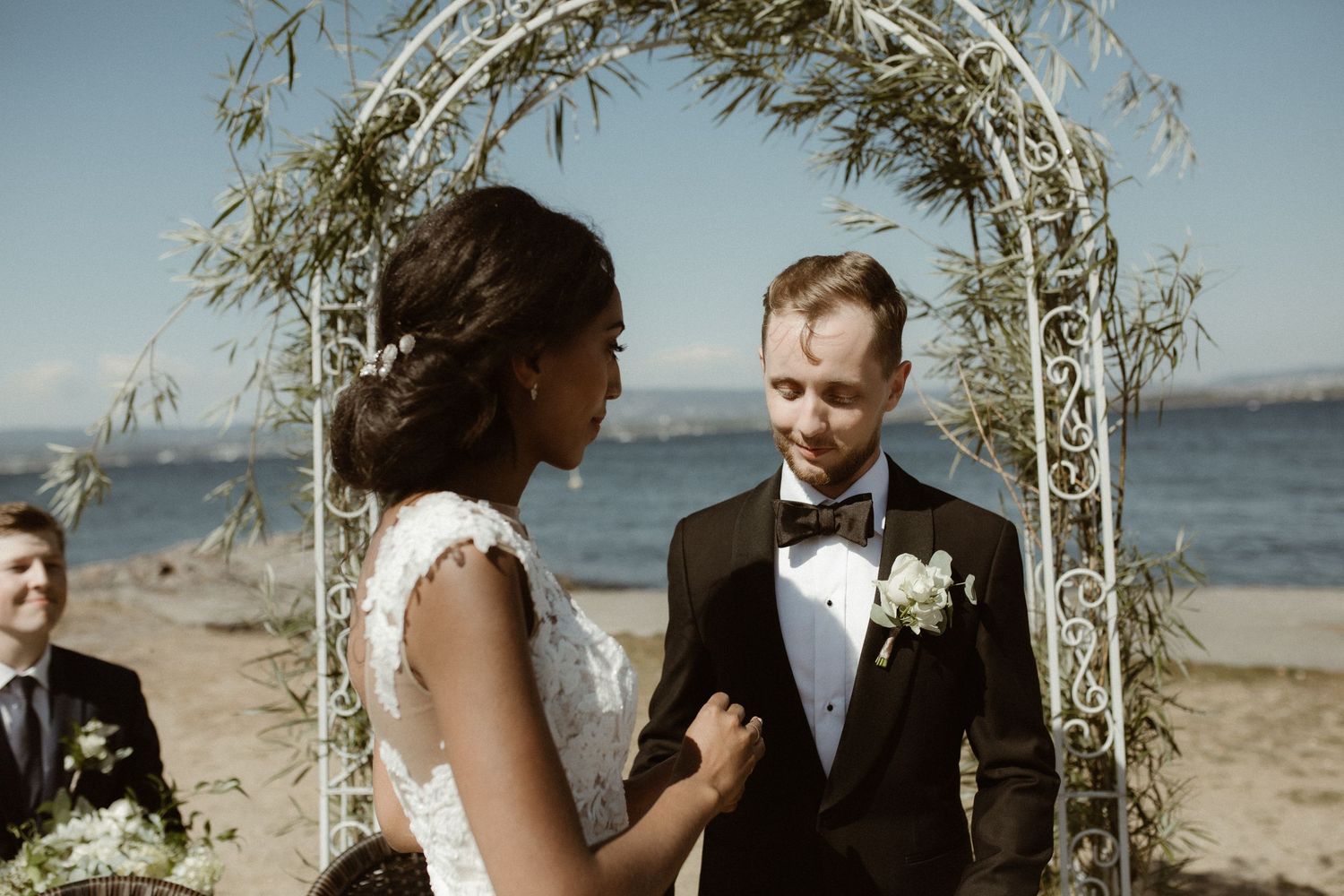 A couple exchanges vows beneath a delicate arch on a beach during their waterfront wedding ceremony.