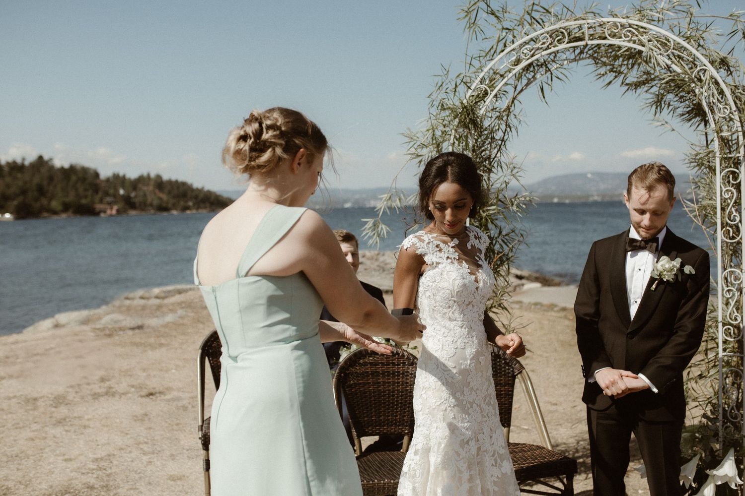 Seaside wedding ceremony under a floral arch with water and mountains in the background.