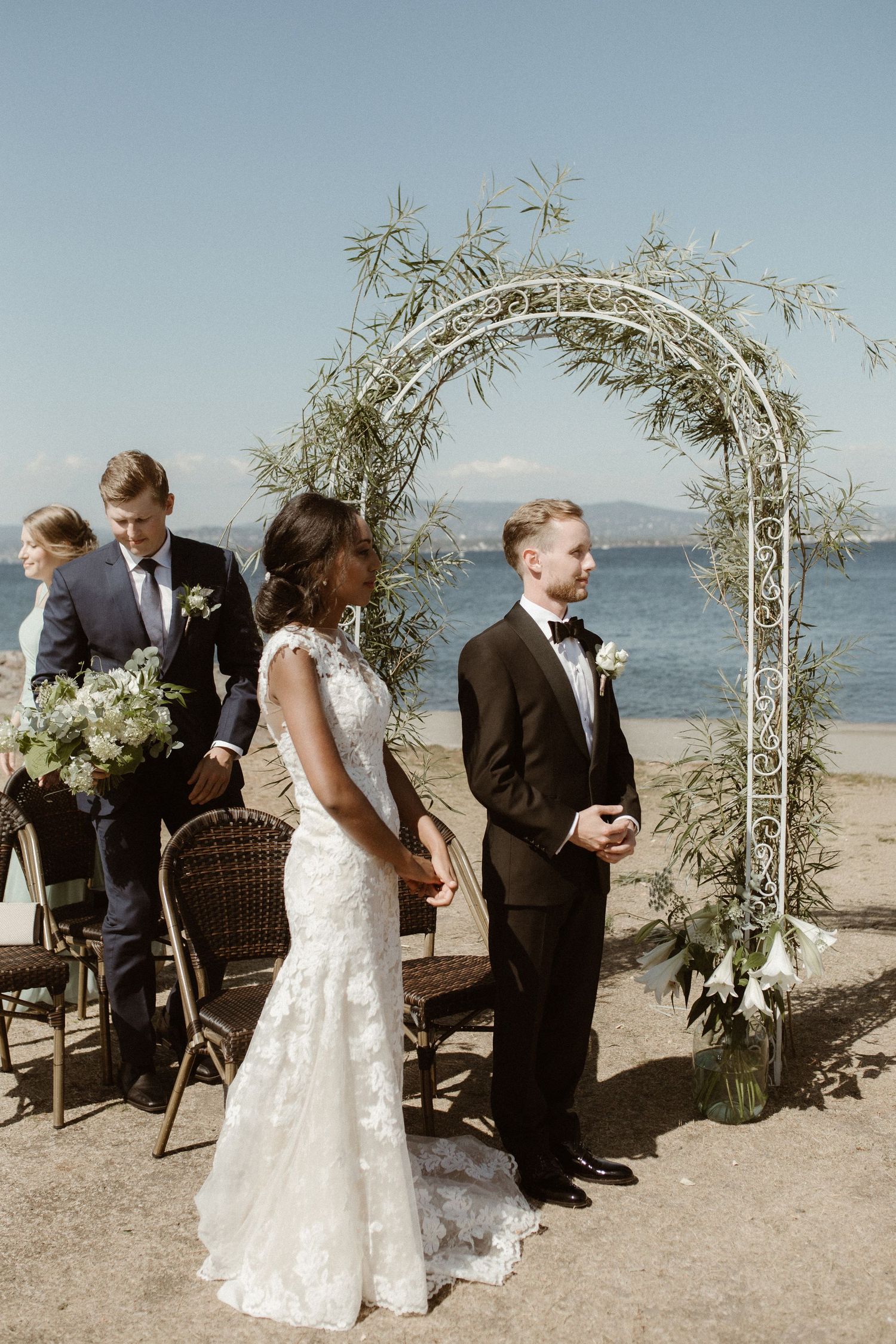 A beachside wedding ceremony under a floral arch with the ocean and mountains in the background.