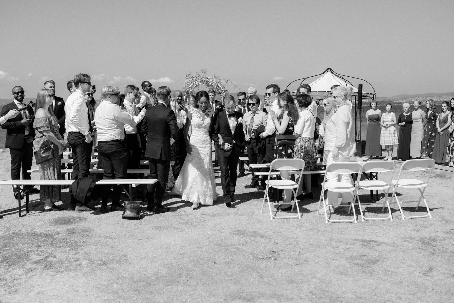 Wedding guests stand during an outdoor ceremony with chairs arranged on a sunny day.
