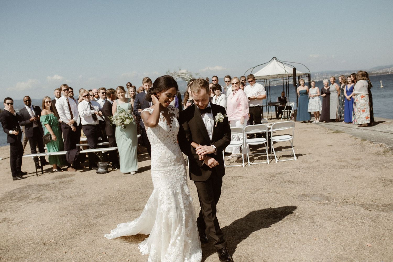 Newlyweds walk together on a beach while wedding guests look on during an outdoor celebration.