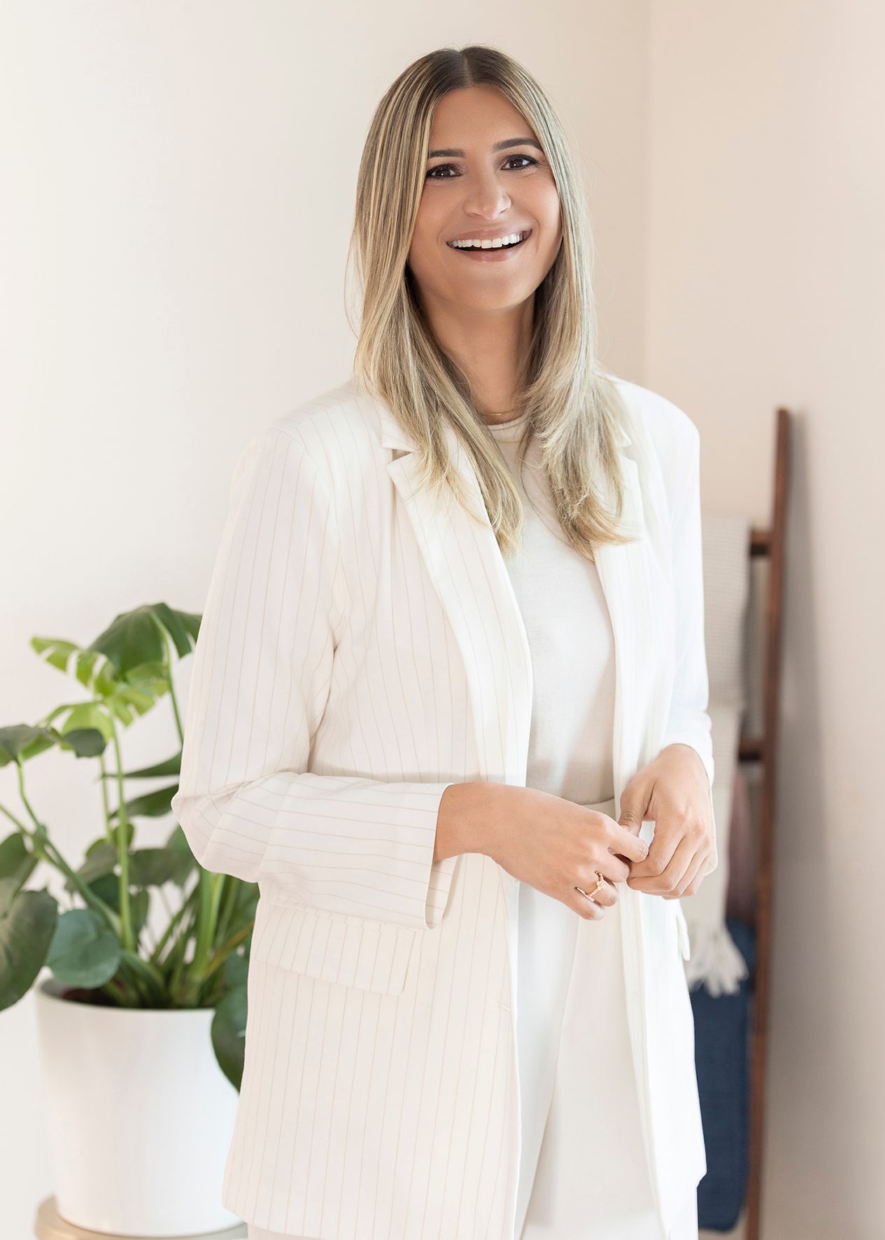 Woman in crisp white blazer outfit smiles warmly against neutral background with decorative indoor plant.