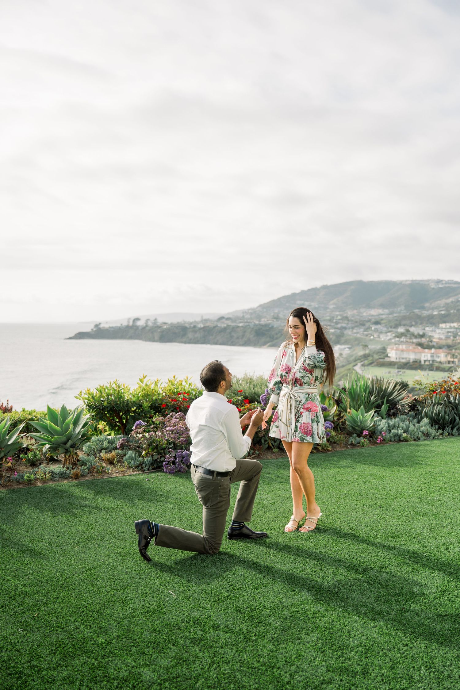 A romantic proposal moment on a scenic cliffside overlooking the ocean with lush landscaping and coastal views.