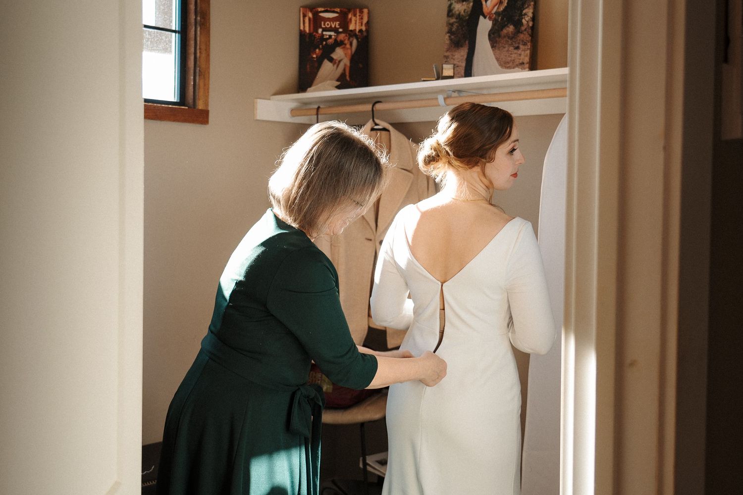 A person in a deep green dress helps adjust the back of an elegant white wedding gown in a sunlit room.