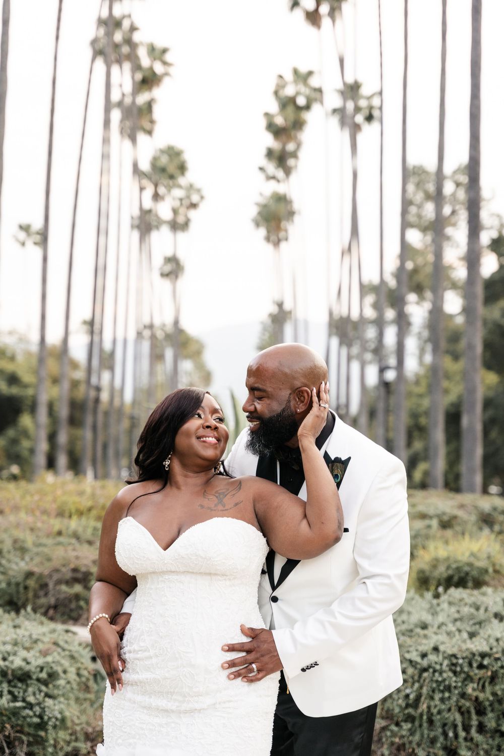 A couple shares a romantic moment in their wedding attire among tall palm trees.