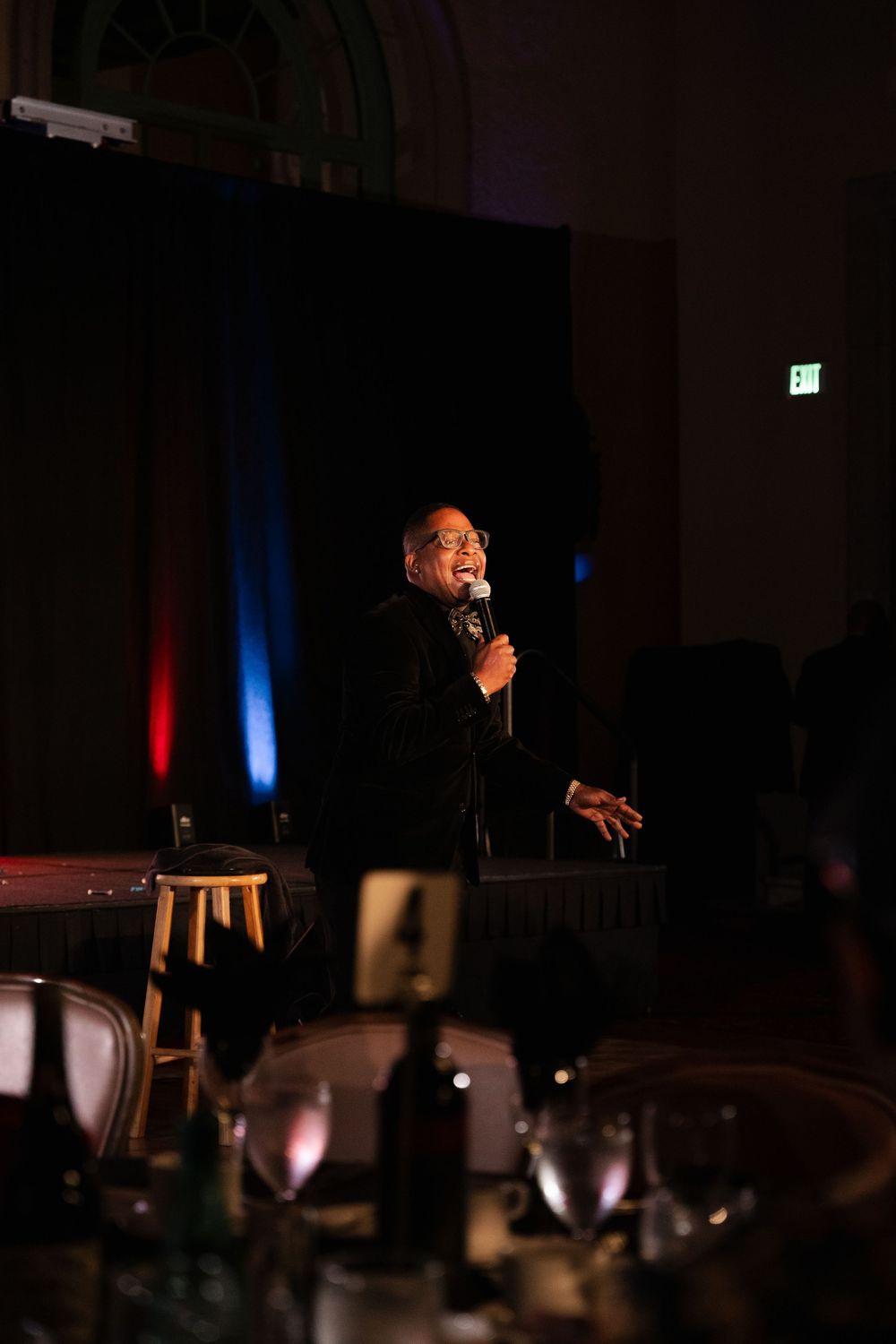 A performer in a black suit speaks into a microphone on a dimly lit stage with red and blue lighting in the background.