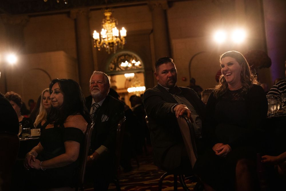 A group of people laugh together in a dimly lit ballroom with elegant chandeliers overhead.