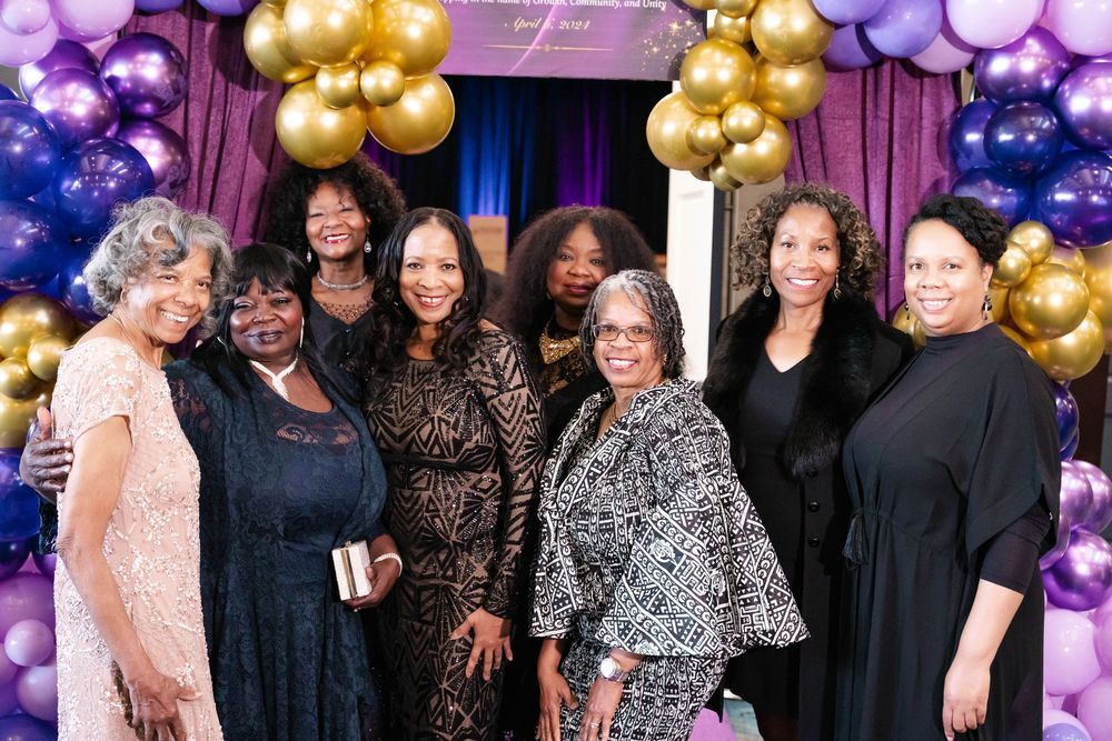 Eight people pose together in front of purple and gold balloon decorations at a celebratory event.
